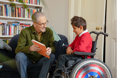 Young boy in a wheelchair, sat next to an older man who is about to read a children's book
